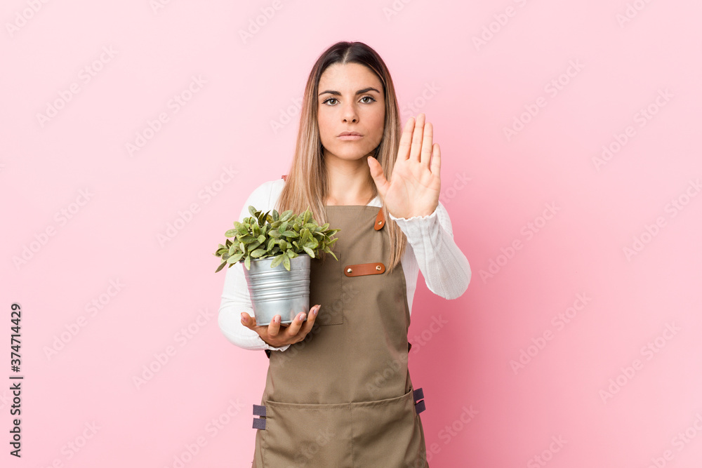 Young gardener woman holding a plant standing with outstretched hand showing stop sign, preventing you. Young gardener woman holding a plant standing with outstretched hand showing stop sign, preventing you.
