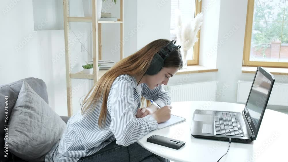 Serious girl student wears headphones, studies with internet teacher, learns language. focused young woman talks to tutor, looks at laptop, writes notes. online education, video call tutoring concept Serious girl student wears headphones, studies with internet teacher, learns language. focused young woman talks to tutor, looks at laptop, writes notes. online education, video call tutoring concept