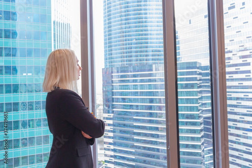 Beautiful caucasian blonde hair woman stands in office. Blurred office buildings are visible through window. Copy space for your text. Theme of Successful women in business.