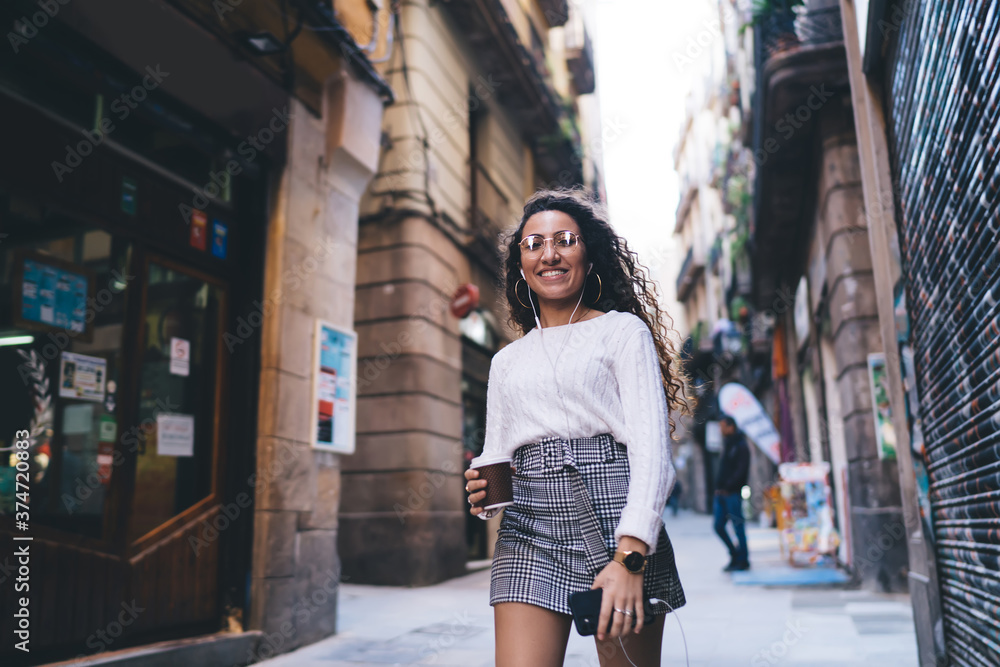 Half length portrait of cheerful female tourist in optical eyewear for vision correction exploring city street during coffee break, joyful hipster girl with smartphone in hand walking during getaway