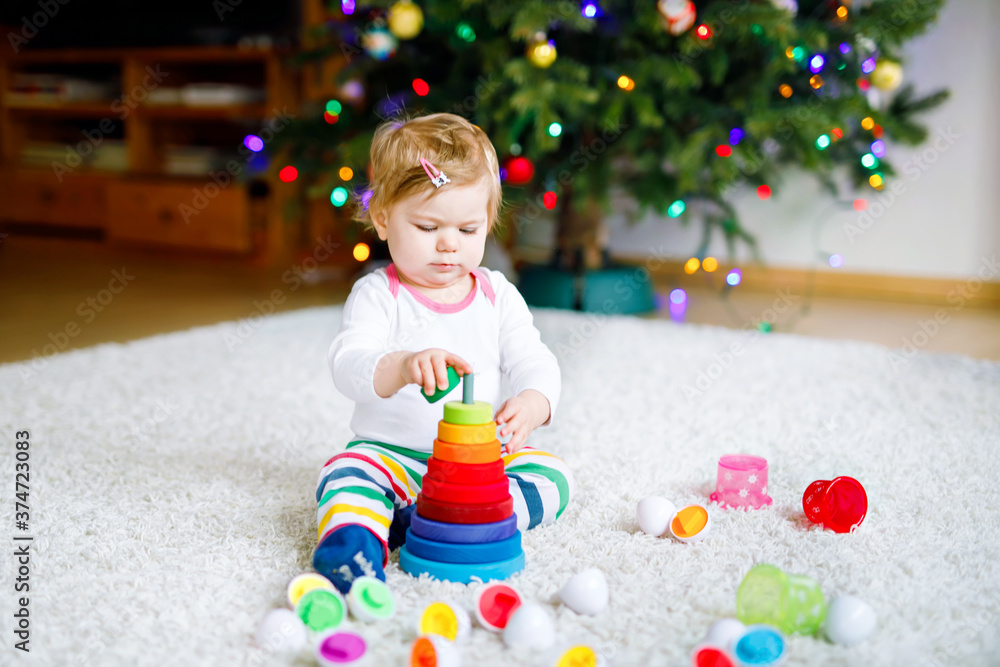 Obraz premium Adorable cute beautiful little baby girl playing with educational toys at home or nursery. Happy healthy child having fun with colorful wooden rainboy toy pyramid. Kid learning different skills.