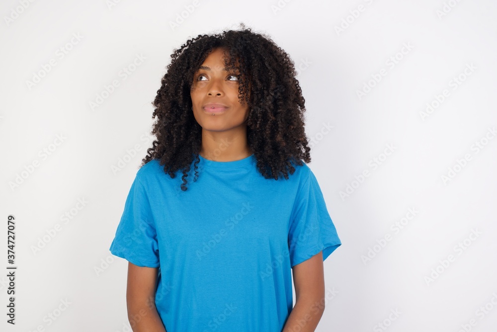 Fototapeta premium Cheerful positive Young african woman with curly hair wearing casual blue shirt over white background, looking aside into empty space thoughtful
