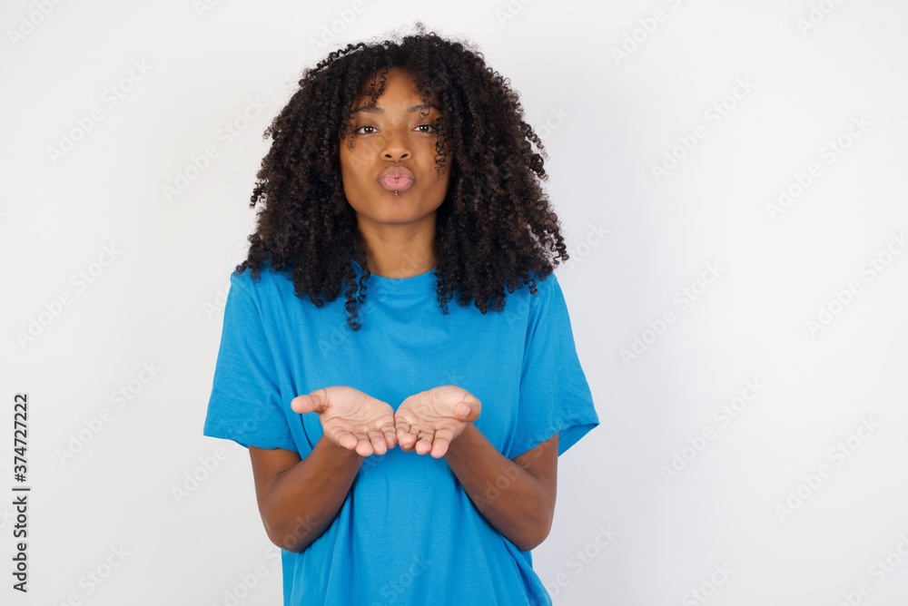 Young african woman with curly hair wearing casual blue shirt over white background sending blow kiss with pout lips and holding palms to send air kiss. Young african woman with curly hair wearing casual blue shirt over white background sending blow kiss with pout lips and holding palms to send air kiss.