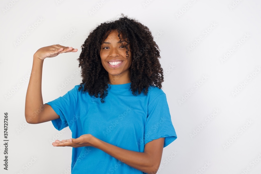 Young african woman with curly hair wearing casual blue shirt over white wall gesturing with hands showing big and large size sign, measure symbol. Smiling looking at the camera. Measuring concept. Young african woman with curly hair wearing casual blue shirt over white wall gesturing with hands showing big and large size sign, measure symbol. Smiling looking at the camera. Measuring concept.