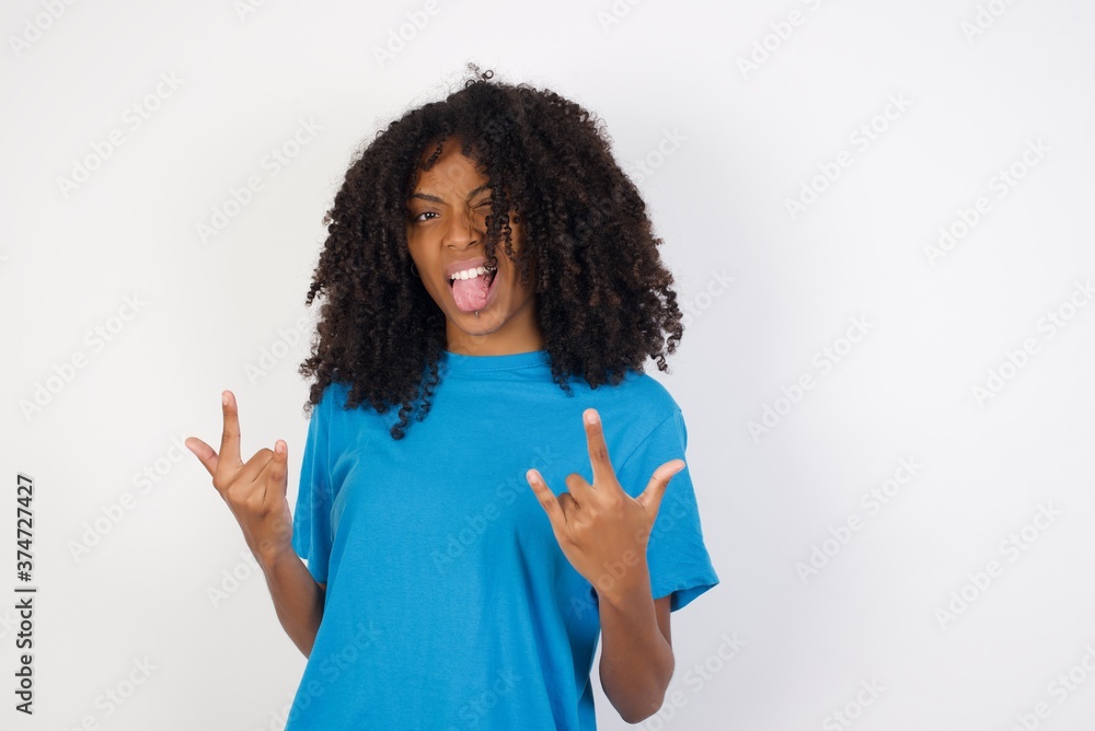 Young african woman with curly hair wearing casual blue shirt over white background posing in studio. showing rock and roll hand gesture Young african woman with curly hair wearing casual blue shirt over white background posing in studio. showing rock and roll hand gesture
