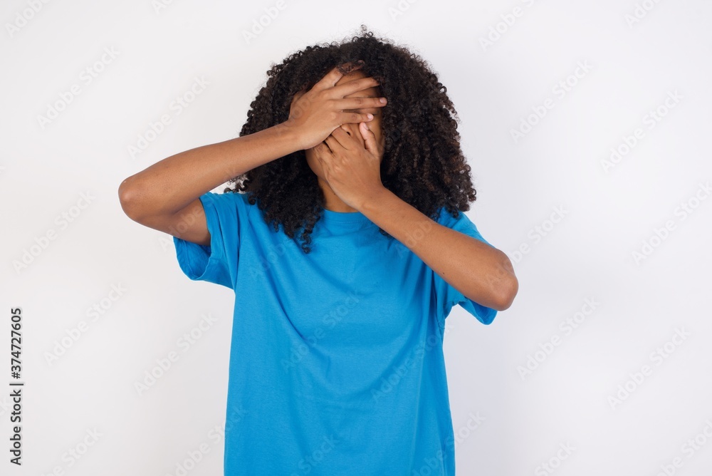 Young african woman with curly hair wearing casual blue shirt over white background Covering eyes and mouth with hands, surprised and shocked. Hiding emotions. Young african woman with curly hair wearing casual blue shirt over white background Covering eyes and mouth with hands, surprised and shocked. Hiding emotions.