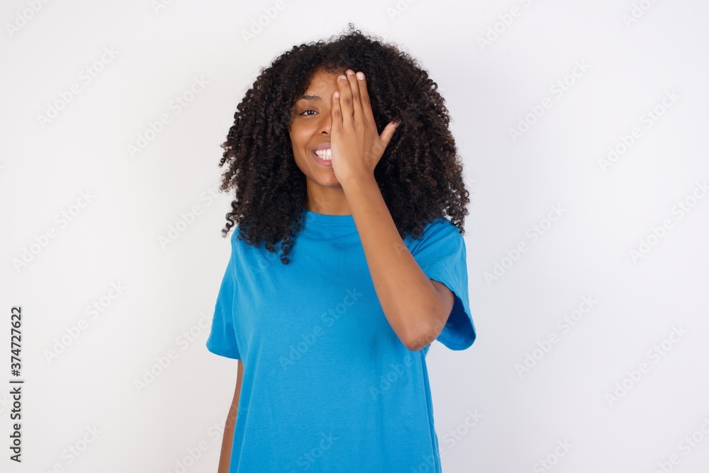 Young african woman with curly hair wearing casual blue shirt over white background covering one eye with her hand, confident smile on face and surprise emotion. Young african woman with curly hair wearing casual blue shirt over white background covering one eye with her hand, confident smile on face and surprise emotion.