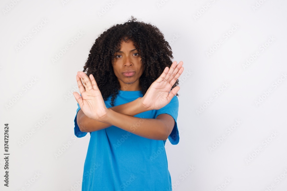 Young african woman with curly hair wearing casual blue shirt over white background has rejection expression crossing arms and palms doing negative sign, angry face. Young african woman with curly hair wearing casual blue shirt over white background has rejection expression crossing arms and palms doing negative sign, angry face.