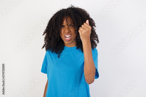 Young african woman with curly hair wearing casual blue shirt over white background being angry and mad raising fist frustrated and furious while shouting with anger. Rage and aggressive concept.