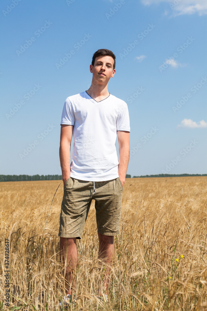 Full length portrait of teenager boy standing on agricultural field with ripe wheat, yellow meadow and blue sky, hands are in pockets of shorts, looking at camera Full length portrait of teenager boy standing on agricultural field with ripe wheat, yellow meadow and blue sky, hands are in pockets of shorts, looking at camera