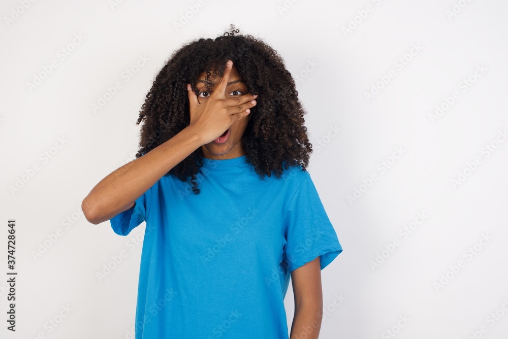 Young african woman with curly hair wearing casual blue shirt peeking in shock covering face and eyes with hand, looking through fingers with embarrassed expression.
