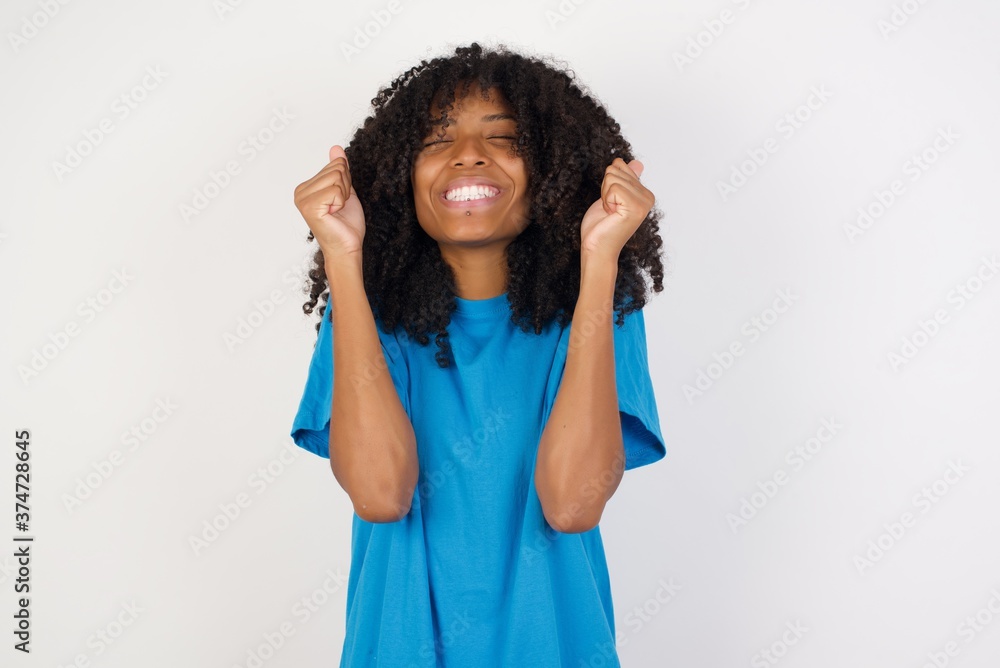 Young african woman with curly hair wearing casual blue shirt being excited for success with raised arms and closed eyes celebrating victory. Winner concept. Young african woman with curly hair wearing casual blue shirt being excited for success with raised arms and closed eyes celebrating victory. Winner concept.