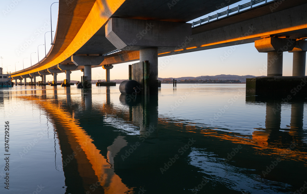 Morning sun strikes side Tauranga Harbour Bridge in golden hue reflected leading lines into calm water below