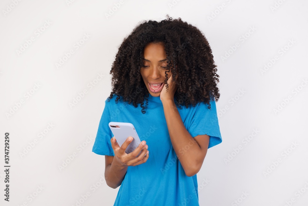 Close-up portrait of Young african woman with curly hair wearing casual blue shirt holding in hands cell reading sms using new app 5g Close-up portrait of Young african woman with curly hair wearing casual blue shirt holding in hands cell reading sms using new app 5g