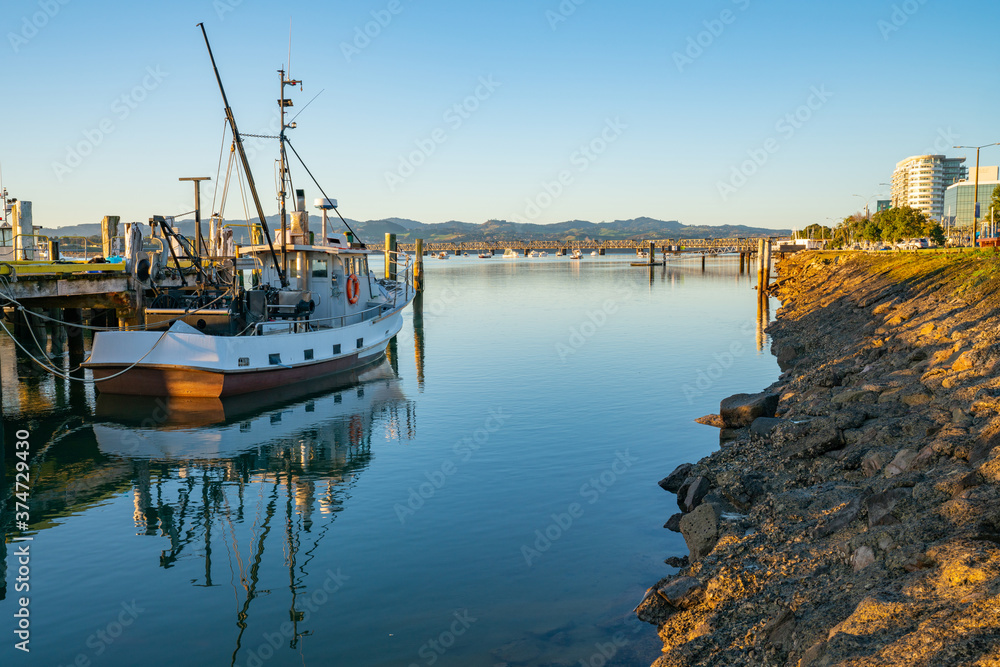 Fototapeta premium Waterfront at Fisherman's Wharf with moored commercial fishing boat and city downtown with railway bridge in distance.
