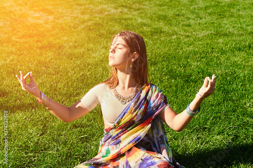 A beautiful young Indian girl in traditional clothes with makeup and jewelry is sitting in a Lotus position on the bright green grass. In the open air under the bright rays of the sun. Indian beauty