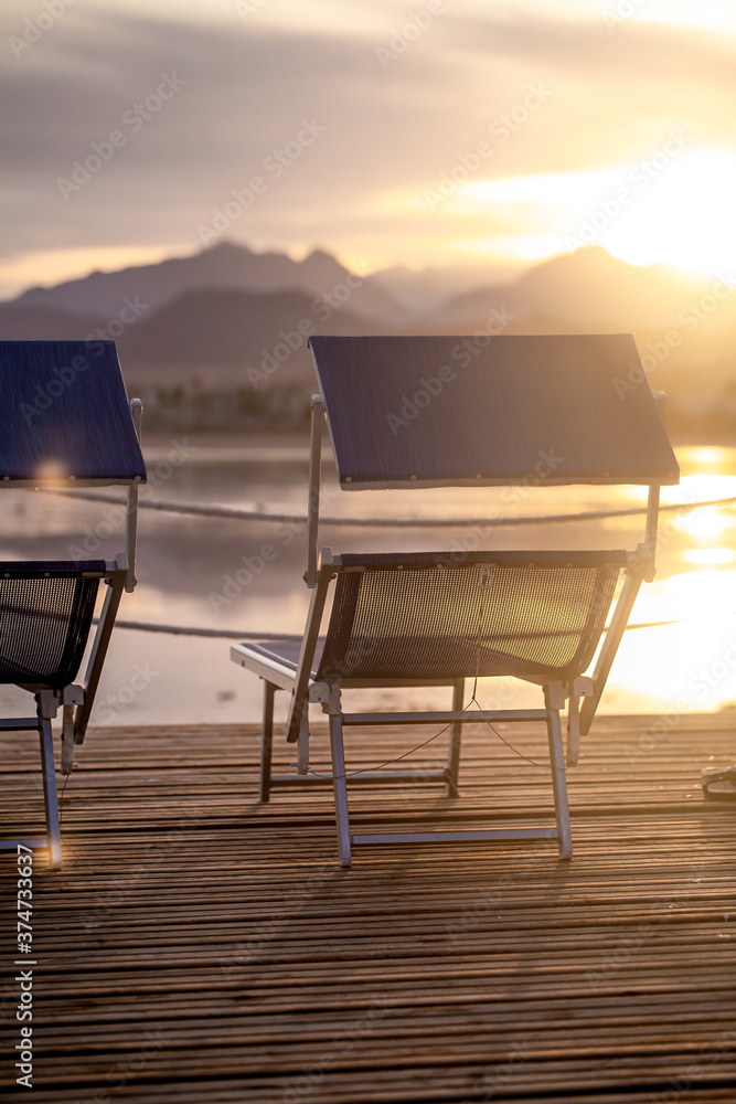 Two empty chairs on a wooden pier at sunset - obrazy, fototapety, plakaty 