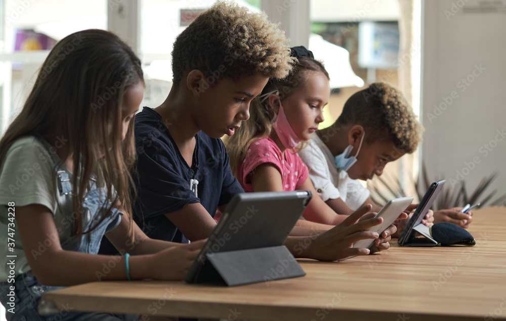 Four diverse kids wear facemasks sit at table use wireless gadgets ignoring each other prefer internet games and virtual communication. Alpha generation and modern technology overuse, phubbing concept Four diverse kids wear facemasks sit at table use wireless gadgets ignoring each other prefer internet games and virtual communication. Alpha generation and modern technology overuse, phubbing concept