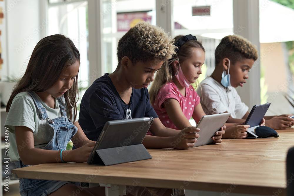 Four diverse kids wear facemasks sit at table use wireless gadgets ignoring each other prefer internet games and virtual communication. Alpha generation and modern technology overuse, phubbing concept Four diverse kids wear facemasks sit at table use wireless gadgets ignoring each other prefer internet games and virtual communication. Alpha generation and modern technology overuse, phubbing concept