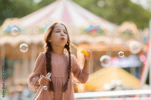 Teenage girl with two pigtails launches bubbles in amusement park near carousel and rejoices