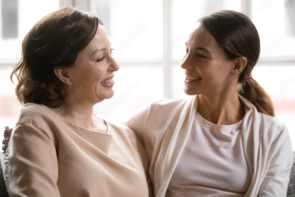 Head shot smiling beautiful brown-haired mature woman communicating with happy grown daughter, sitting together on couch indoors. Cheerful female family sharing news, enjoying pleasant conversation. Head shot smiling beautiful brown-haired mature woman communicating with happy grown daughter, sitting together on couch indoors. Cheerful female family sharing news, enjoying pleasant conversation.