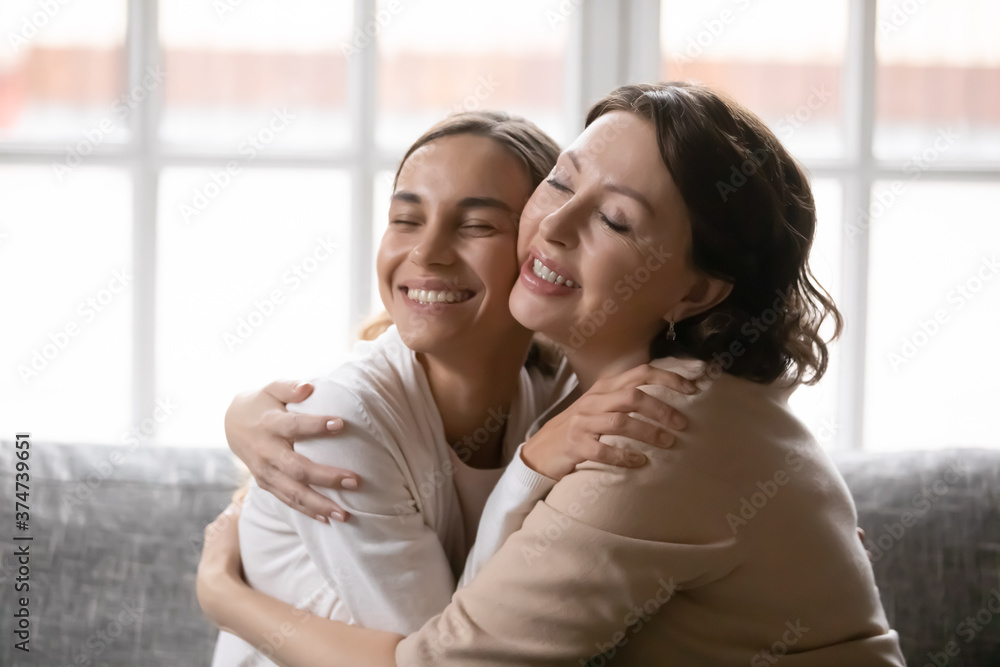 Fototapeta premium Happy tender middle aged senior woman cuddling smiling grown up daughter, relaxing together on comfortable couch at home. Affectionate multigenerational female family enjoying sweet moment indoors.