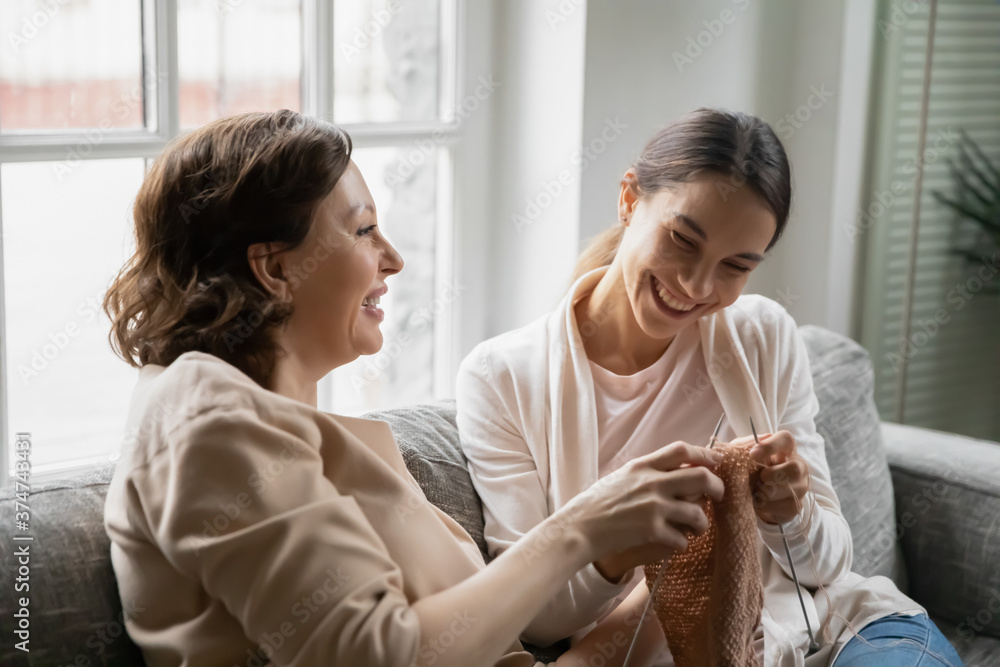 Φωτογραφία Happy middle aged 60s woman resting on comfortable sofa, teaching grown up smiling mixed race daughter knitting warm scarf with wooden threads indoors, active family hobby weekend pastime at home
