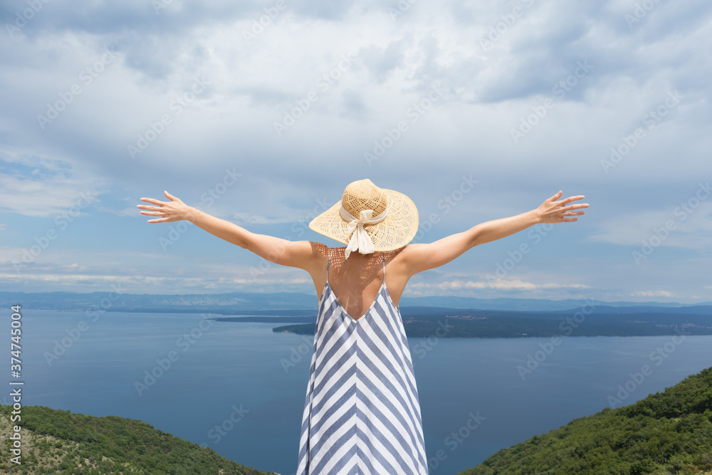 Φωτογραφία Rear view of young woman wearing striped summer dress and straw hat standing in super bloom of wildflowers, relaxing with hands up to the sky, enjoing beautiful view of Adriatic sea nature, Croatia