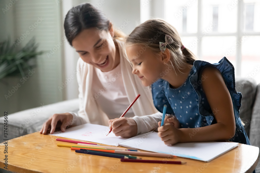 Smiling millennial mother or older sister enjoying drawing pictures with small child girl in paper album, spending weekend hobby time together at home, sitting at table, creative skills development. Smiling millennial mother or older sister enjoying drawing pictures with small child girl in paper album, spending weekend hobby time together at home, sitting at table, creative skills development.