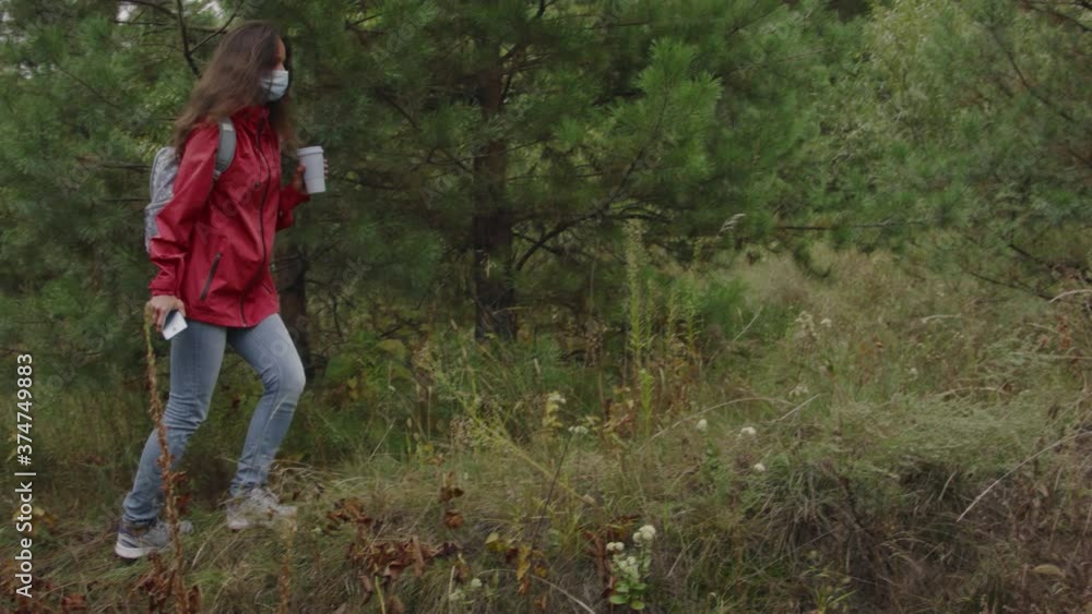 Young woman in a coronavirus mask walking through the forest with an eco cup of coffee and a smartphone in her hands on a cloudy autumn day. Young woman in a coronavirus mask walking through the forest with an eco cup of coffee and a smartphone in her hands on a cloudy autumn day.