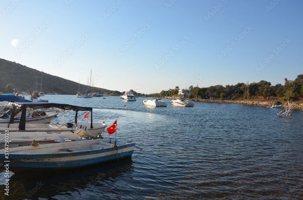 View from the shore of boats and sailboats moored in a bay of the Aegean Sea. Panoramic view of the entire bay. Boats with Turkish flag moored to the pier. The hill rising at the end of the bay. 
