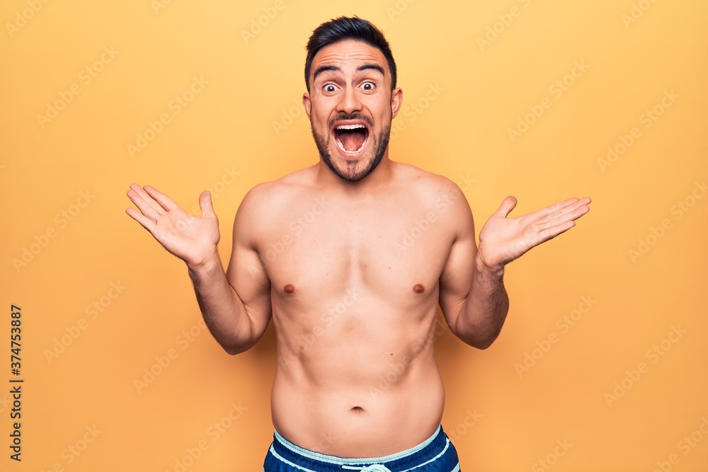 Young handsome man with beard wearing sleeveless t-shirt standing over yellow background celebrating victory with happy smile and winner expression with raised hands