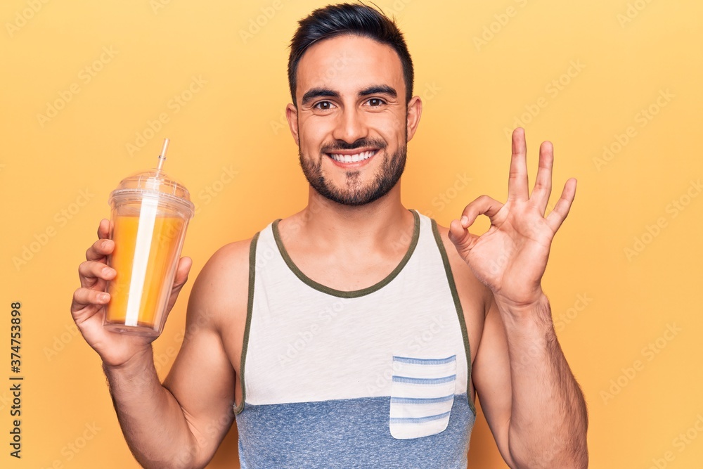 Handsome man with beard wearing sleeveless t-shirt drinking glass of healthy orange juice doing ok sign with fingers, smiling friendly gesturing excellent symbol Handsome man with beard wearing sleeveless t-shirt drinking glass of healthy orange juice doing ok sign with fingers, smiling friendly gesturing excellent symbol