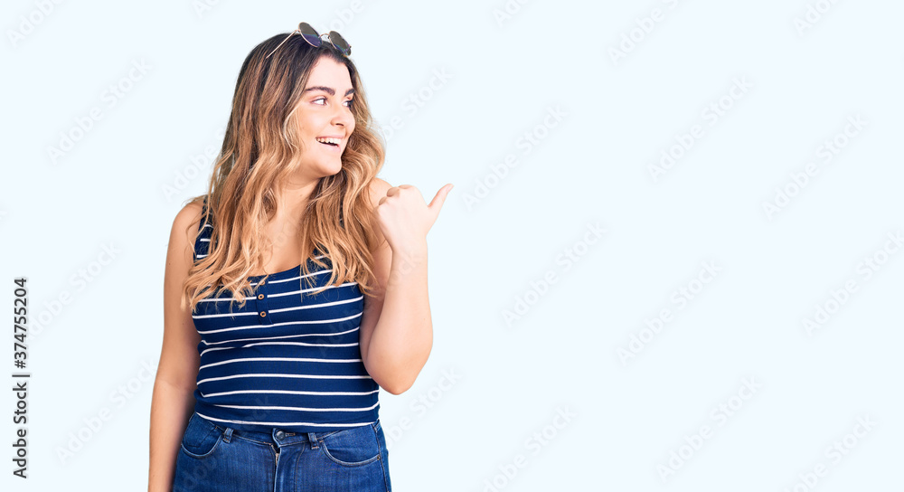 Fototapeta premium Young caucasian woman wearing casual clothes and sunglasses smiling with happy face looking and pointing to the side with thumb up.