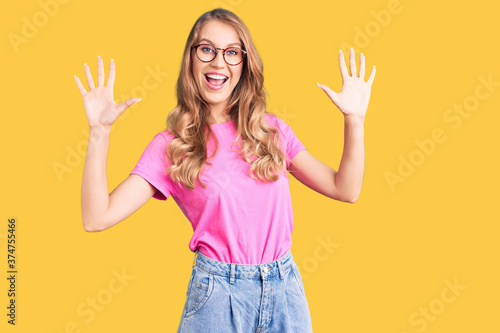 Young beautiful caucasian woman with blond hair wearing casual clothes and glasses showing and pointing up with fingers number ten while smiling confident and happy.