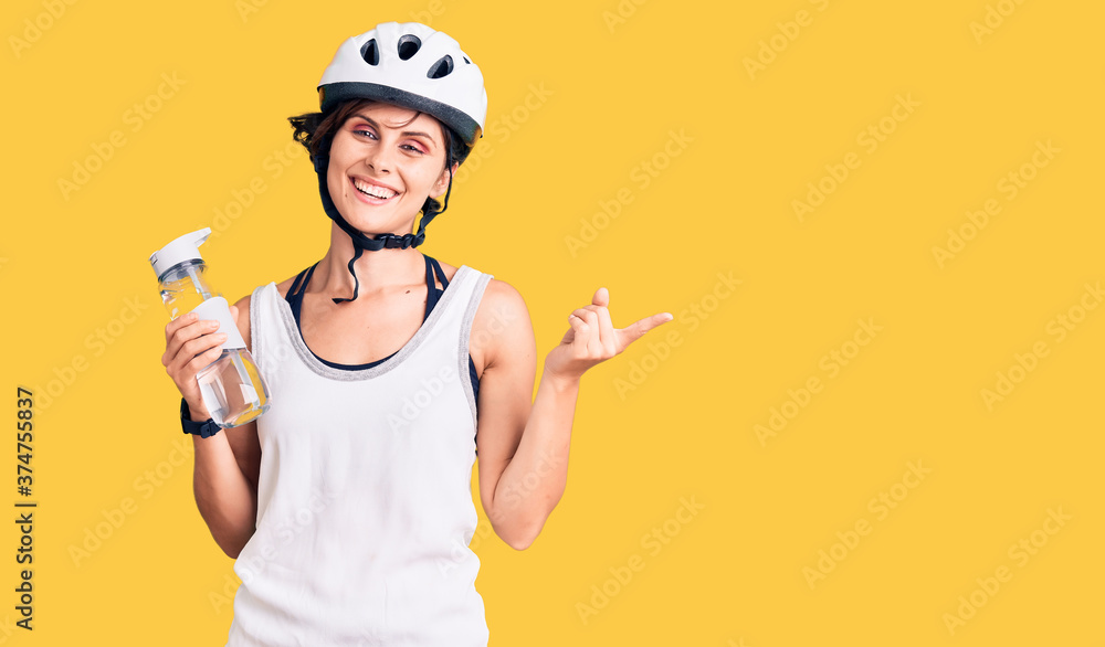 Beautiful young woman with short hair wearing bike helmet and holding water bottle smiling happy pointing with hand and finger to the side Beautiful young woman with short hair wearing bike helmet and holding water bottle smiling happy pointing with hand and finger to the side