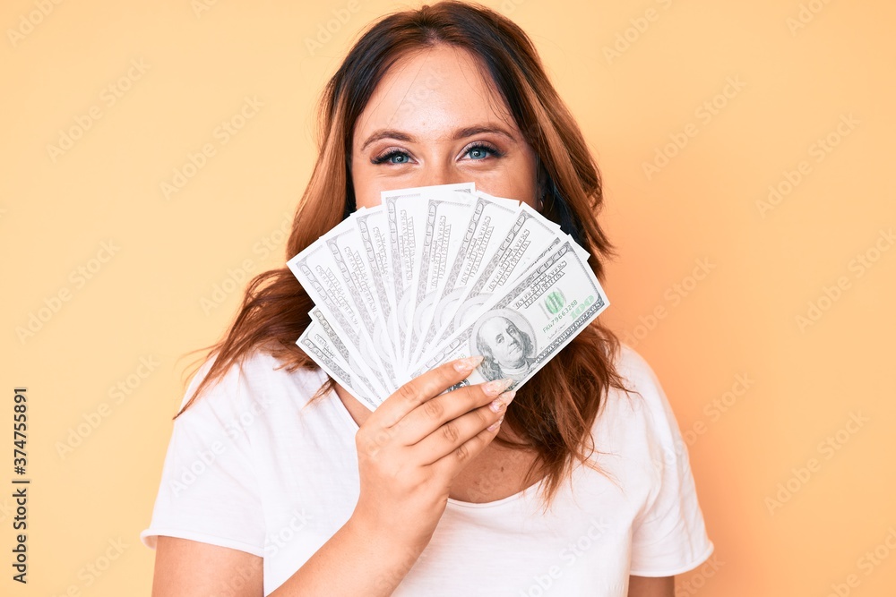 Young beautiful caucasian woman holding dollars looking positive and happy standing and smiling with a confident smile showing teeth Young beautiful caucasian woman holding dollars looking positive and happy standing and smiling with a confident smile showing teeth