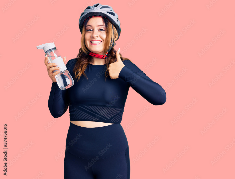 Young beautiful caucasian woman wearing bike helmet and holding water bottle smiling happy and positive, thumb up doing excellent and approval sign Young beautiful caucasian woman wearing bike helmet and holding water bottle smiling happy and positive, thumb up doing excellent and approval sign
