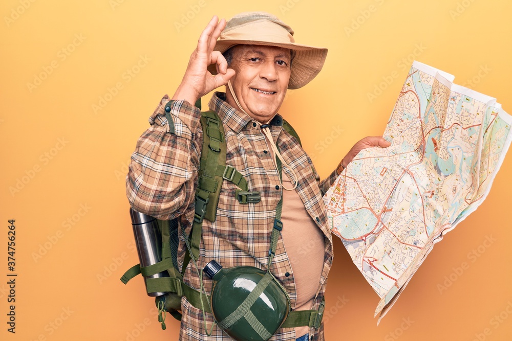 Senior man with grey hair wearing hiker backpack holding map doing ok sign with fingers, smiling friendly gesturing excellent symbol