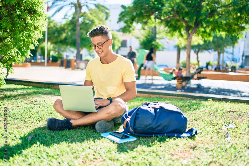 Obraz premium Young hispanic student smiling happy using laptop sitting on the grass at the university campus