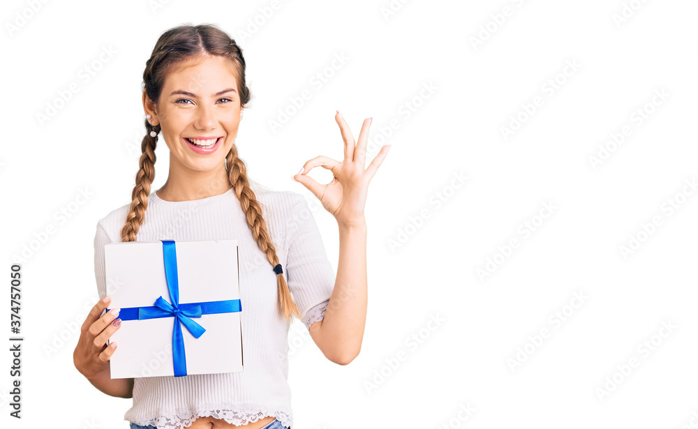 Beautiful caucasian woman with blonde hair holding gift doing ok sign with fingers, smiling friendly gesturing excellent symbol Beautiful caucasian woman with blonde hair holding gift doing ok sign with fingers, smiling friendly gesturing excellent symbol