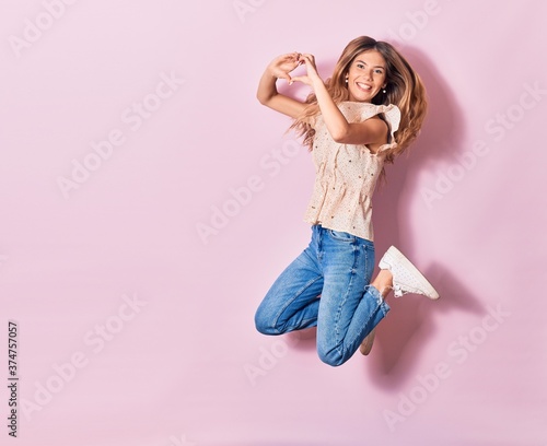 Young beautiful woman wearing casual clothes smiling happy. Jumping with smile on face doing heart shape with fingers over isolated pink background