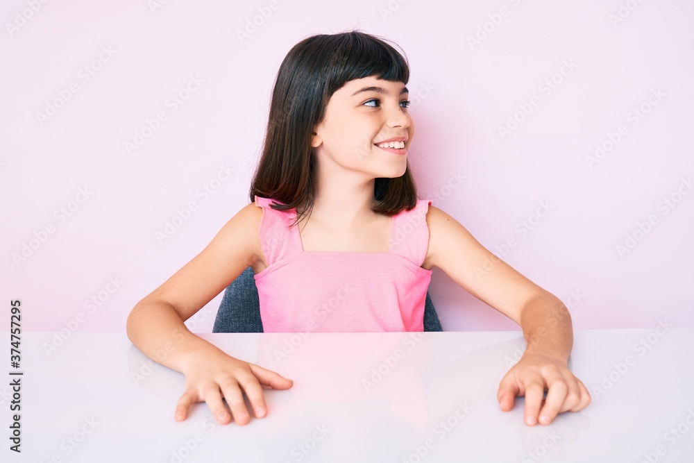 Young little girl with bang wearing casual clothes sitting on the table looking away to side with smile on face, natural expression. laughing confident. Young little girl with bang wearing casual clothes sitting on the table looking away to side with smile on face, natural expression. laughing confident.