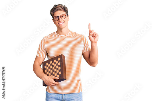 Young handsome man holding chess surprised with an idea or question pointing finger with happy face, number one