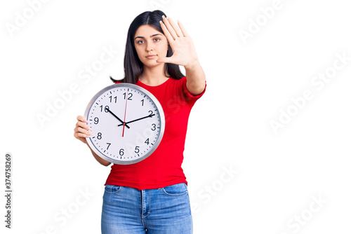 Young beautiful girl holding clock with open hand doing stop sign with serious and confident expression, defense gesture