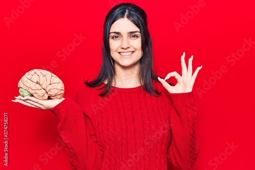 Young beautiful girl holding brain doing ok sign with fingers, smiling friendly gesturing excellent symbol