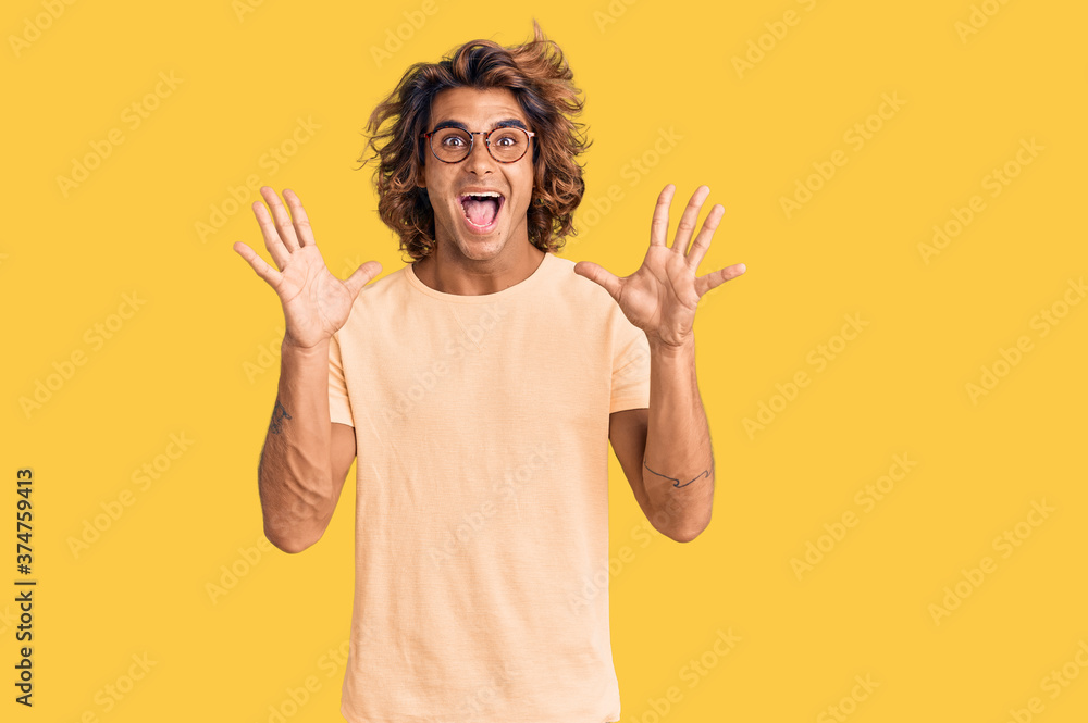 Young hispanic man wearing casual clothes and glasses celebrating crazy and amazed for success with arms raised and open eyes screaming excited. winner concept Young hispanic man wearing casual clothes and glasses celebrating crazy and amazed for success with arms raised and open eyes screaming excited. winner concept
