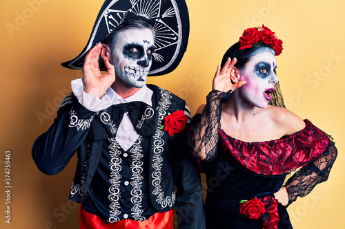 Young couple wearing mexican day of the dead costume over yellow smiling with hand over ear listening an hearing to rumor or gossip. deafness concept.