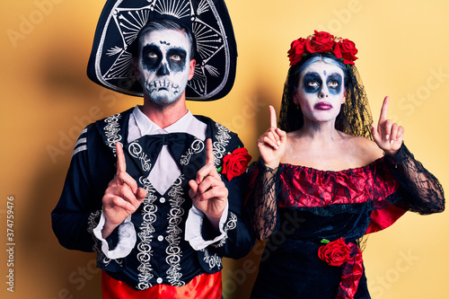 Young couple wearing mexican day of the dead costume over yellow pointing up looking sad and upset, indicating direction with fingers, unhappy and depressed.