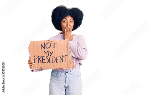 Young african american girl holding not my president protest banner covering mouth with hand, shocked and afraid for mistake. surprised expression © Krakenimages.com